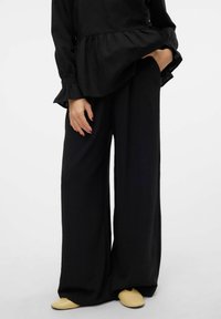 Czarny top peplum z długimi rękawami, zestawiony z szerokimi czarnymi spodniami. Żółte buty wsuwane dopełniają strój. Miękka tekstura, prosty design.