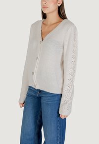 Cárdigan beige claro con cuello en V, textura acanalada y patrones de punto decorativos en las mangas. Presenta cinco botones en la parte delantera.