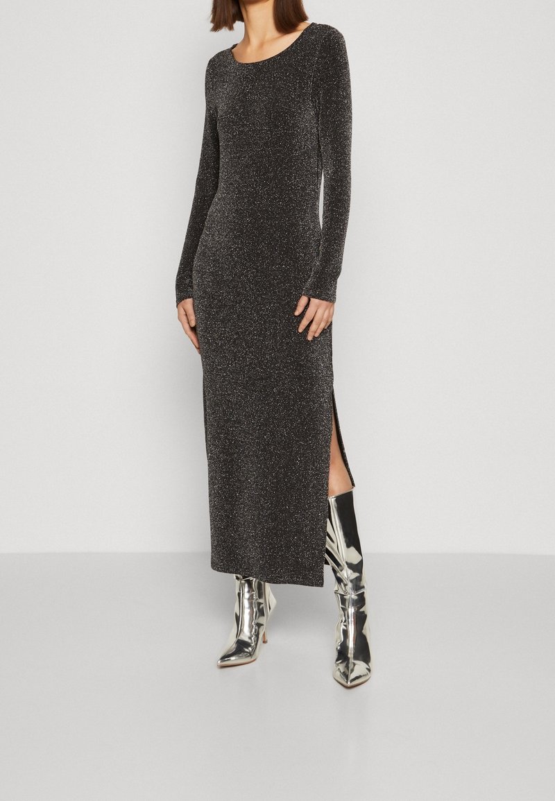 Femme portant une robe noire scintillante à manches longues avec une fente latérale, associée à des bottes à talons hauts argentées brillantes sur un fond uni.