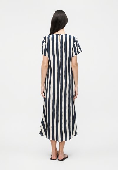 Γυναίκα με μακριά σκουρόχρωμα μαλλιά, που φοράει μια midi ριγέ φόρεμα σε ναυτικό μπλε και λευκό χρώμα με κάθετες ρίγες και μαύρα επίπεδα σανδάλια, στέκεται με την πλάτη γυρισμένη σε ένα απλό φόντο.