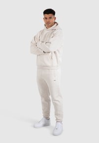 Felpa grigia chiara con cappuccio e pantaloni coordinati in tessuto morbido, dotati di tasca frontale, maniche elastiche e tasche sui lati. Sneakers bianche.