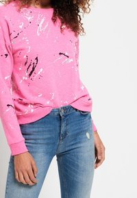 Rosa sweatshirt med vita och svarta splash-mönster, rund halsringning och ribbade muddar, tillsammans med blå slitna jeans.
