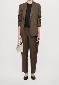 Kvinna iklädd brun dubbelknäppt kavaj och byxor, svart genomskinlig topp, svarta platta skor, håller en liten vit handväska framför en vit bakgrund.