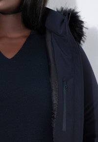 Chaqueta de plumón azul marino con capucha de piel sintética desmontable, que cuenta con un bolsillo en el pecho con cremallera y un forro interior acolchado. Debajo lleva un jersey de punto oscuro con escote en V.