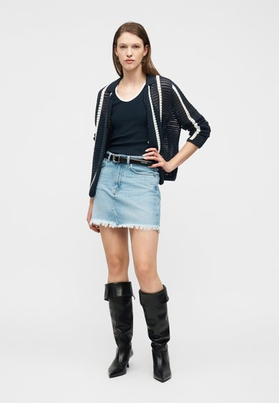 Γυναίκα που φοράει μαύρο πλεκτό ζακέτα με λευκές ρίγες, μαύρο αμάνικο τοπ, φθαρμένη ανοιχτόχρωμη τζιν φούστα, μαύρη ζώνη και μαύρες μπότες μέχρι το γόνατο.
