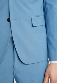 Chaqueta de traje azul claro en una tela suave, con un solo botón negro, solapas de muesca y un bolsillo de solapa a la izquierda.