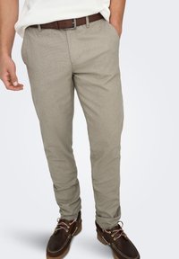 Pantalon beige clair pour homme avec un tissage texturé, coupe slim, poches latérales, accompagné de chaussures marron et d'une ceinture marron.
