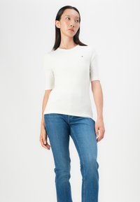White ribbed short-sleeve top with a crew neckline, paired with blue straight-leg jeans. Features a small logo on the chest.