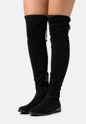 Bottes plates en daim noir montant au-dessus du genou avec fermeture éclair latérale et lien réglable en haut, portées sur des jambes nues sur un fond uni.