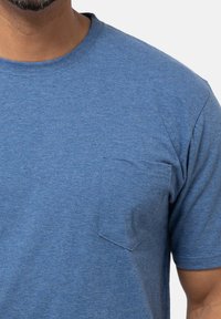 Uomo che indossa una maglietta blu a maniche corte con colletto tondo e una piccola tasca sul lato sinistro.