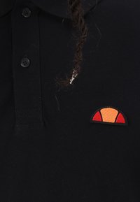 Melns polo krekls ar trim pogām, kuram ir teksturēts oranžs un sarkans logo uzlīme kreisajā pusē netālu no apkakles.