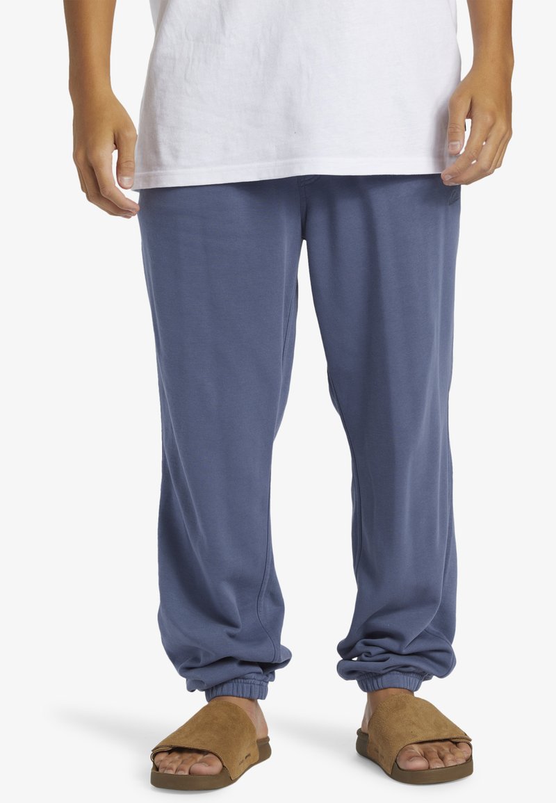 Blå joggers i bomullsblandning med elastiska muddar vid anklarna; matchat med bruna mockasandaler. Avslappnad, komfortabel passform.