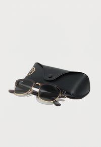 Μαύρα και χρυσά γυαλιά ηλίου Ray-Ban με σκοτεινούς φακούς που ακουμπούν μπροστά σε μια μαύρη θήκη από δέρμα με υφή, με κουμπί κλεισίματος και χρυσό λογότυπο.