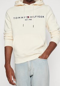 Kreemivärvi kapuutsiga pusa pehmest kangast, millel on ees küljes kangaroo-tasku, tagumine kapuutsiköis ning tikkitud kaubamärgi logo tumesinisena ja punasena.