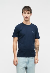 Navy blue cotton T-shirt with a crew neck, short sleeves, and a small embroidered logo on the left chest, paired with light jeans.