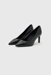 Paire de chaussures à talons hauts en cuir noir à bout pointu avec talons fins sur un fond gris clair.