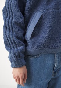 Chaqueta azul de forro polar con tejido texturizado, con tres franjas en la manga y un bolsillo lateral en tejido liso. Diseño cropped.