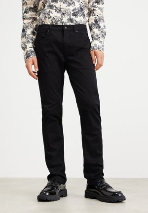 Homme portant un pantalon slim noir, des chaussures de ville noires et brillantes, et une chemise blanche à manches longues avec un imprimé noir détaillé, debout sur un sol clair.