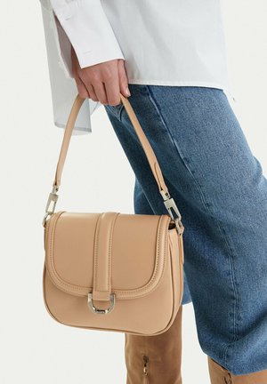 Persona che indossa jeans blu e camicia bianca, che tiene una borsa in pelle color cuoio con fibbia argentata e tracolla regolabile.
