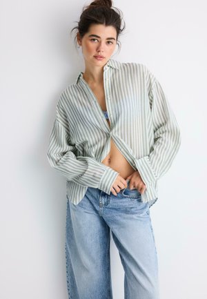 Mladá žena s tmavým drdolom, ktorá nosí voľnú zeleno-bielu pruhovanú košeľu cez modrú podprsenku a svetlo modré rifle s vysokým pásom.