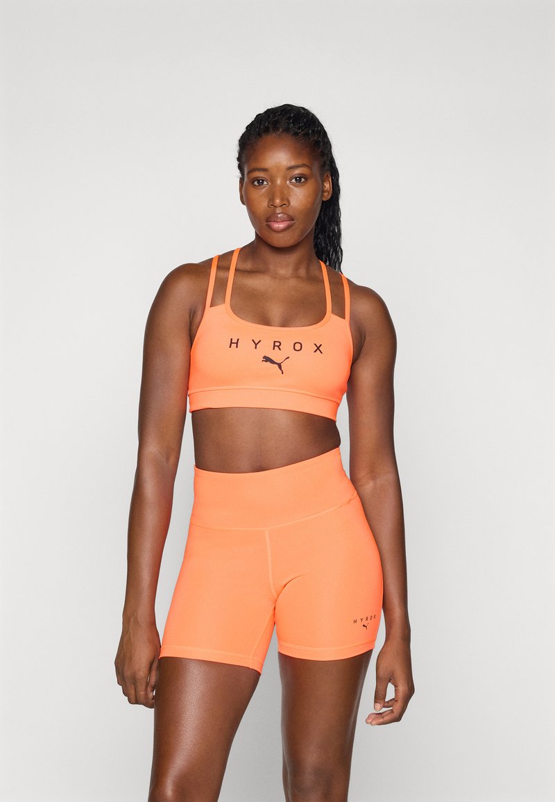 Oransje sport-bh med svart "HYROX" logo og matchende høyt liv shorts. Materialet ser ut til å være elastisk med en glatt tekstur.