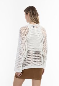 Witte gebreide trui met een open weefpatroon, losse pasvorm en geribde manchetten, gecombineerd met een bruine rok. Zichtbaar detail van de blootgestelde naden.