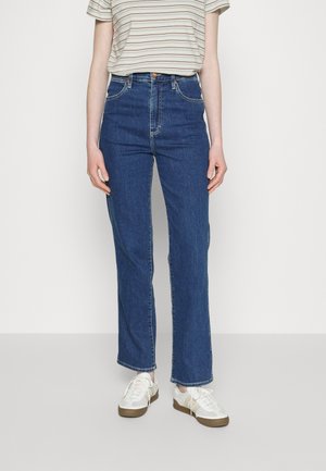 Jean taille haute bleu foncé, coupe droite, avec cinq poches et coutures contrastées. Associé à un haut à rayures à manches courtes et des baskets.