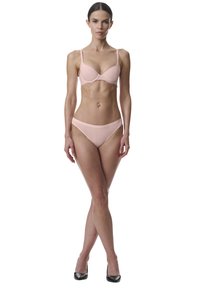 Femme debout, les jambes croisées, portant un soutien-gorge et une culotte rose pâle avec des talons hauts noirs sur fond blanc.