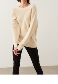 Suéter de punto color crema con un corte holgado, escote amplio y mangas largas, combinado con pantalones ajustados negros. Textura suave.