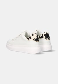 Las zapatillas blancas cuentan con una parte superior de cuero liso y un talón texturizado con un patrón en blanco y negro. El diseño incluye ojales metálicos y una suela gruesa.