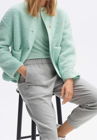 Giacca in pile texturizzato verde menta con due tasche frontali e chiusura a bottoni, abbinata a una maglietta verde chiaro e pantaloni grigi a sigaretta.