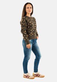 Giovane donna in piedi che indossa un maglione marrone con stampa leopardata, jeans blu e scarpe da ginnastica beige con dettagli in bordeaux e giallo.
