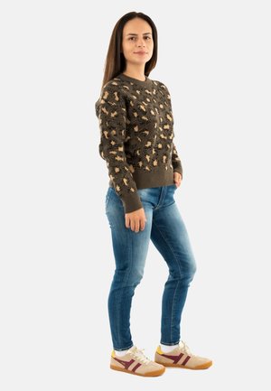 Jeune femme debout portant un pull en imprimé léopard marron, un jean bleu et des baskets beige avec des accents bordeaux et jaunes.