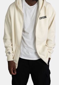 Felpa con zip di colore crema, dotata di tasca frontale, testo "CRYSTAL" nero e logo a forma di diamante sul polsino sinistro. Realizzata in materiale morbido e strutturato.