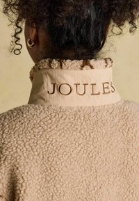 Curly-haired person wearing beige fleece jacket with "JOULES" embroidered on the back collar and gold hoop earring visible.