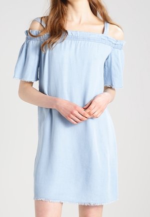 Kvinde iført lyseblå off-shoulder kjole med korte ærmer og frynset kant, stående mod en ensfarvet baggrund.