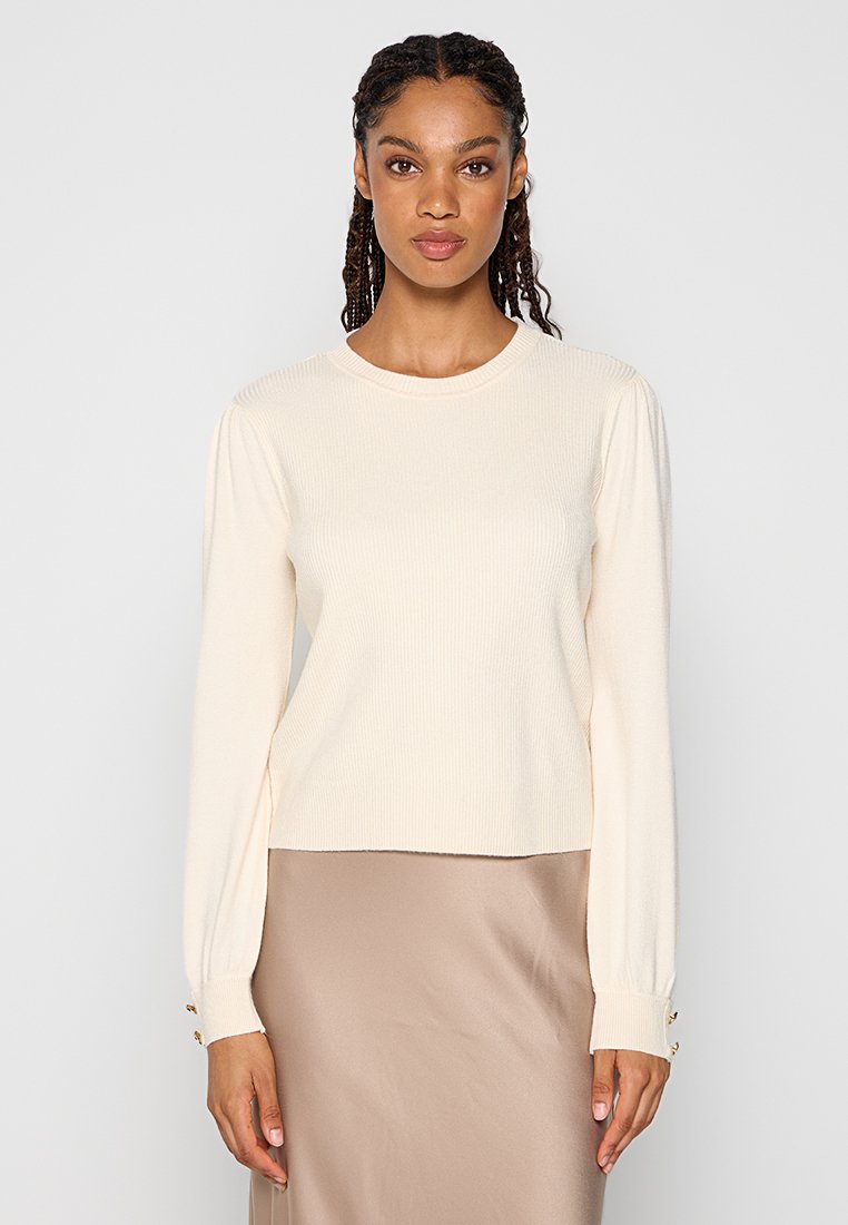 Cremefarbener, gerippter langärmeliger Pullover mit Rundhalsausschnitt und leicht puffigen Ärmeln. Mit Knopfverzierungen an den Ärmelsäumen.