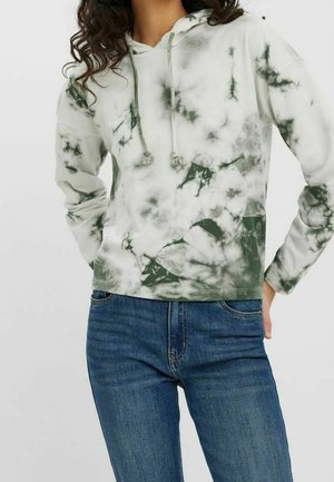 Femme portant un sweat à capuche tie-dye vert et blanc avec poche avant et jean bleu, debout les mains dans les poches.