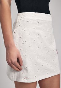 Jupe mini blanche brodée avec un motif floral en œillets et une texture lisse, présentant une taille ajustée et un ourlet droit.