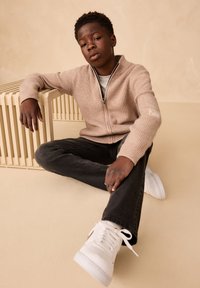 Suéter beige tejido con cremallera y textura acanalada, superpuesto sobre una camisa gris. Jeans negros y zapatillas blancas completan el conjunto.