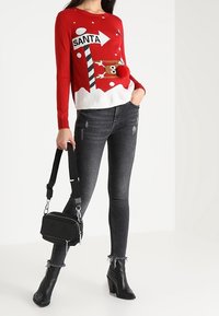 Pull rouge avec un imprimé "Santa" et des flocons de neige blancs, associé à un jean skinny noir et un petit sac à main noir. Bottines à talons noires.