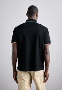 Homme portant un polo noir à manches courtes avec bordure colorée et pantalon beige, debout de dos devant un fond uni.