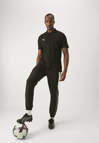 Svart pikétröja med korta ärmar och en vit logotyp, tillsammans med svarta koniska joggers med vita sidodetaljer. Står på en fotboll.