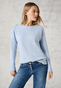 Ljusblå stickad tröja med rundad halsringning och tre knappar på vänster axel, tillsammans med blå jeans med hög midja som har knapplist.