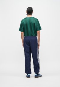 Πράσινο σατέν μπλουζάκι με κοντά μανίκια συνδυασμένο με ναυτικά φόρμας με λευκές ρίγες στα πλάγια. Το μοντέλο φοράει μπλε αθλητικά παπούτσια, εμφανιζόμενο από πίσω.