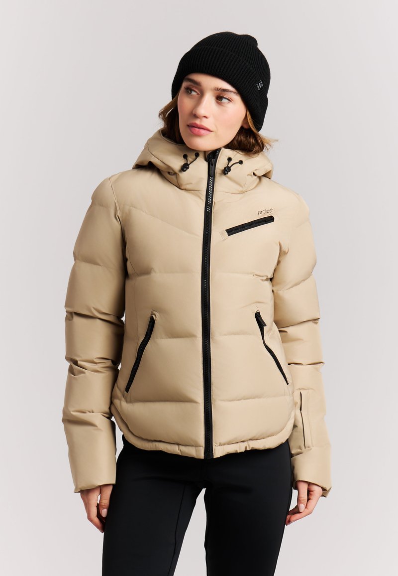Woman wearing a beige puffer jacket with black zippers and a black beanie, looking slightly to her left against a plain background.
