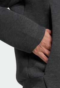 Manche d'un hoodie en tissu gris foncé avec une finition texturée. La main est placée dans une poche latérale, mettant en valeur la couture et les détails de la poche.