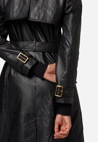 Zwarte leren trenchcoat met een ceintuur in de taille, voorzien van geribde zwarte manchetten en goudkleurige gespen, met een strakke, gestructureerde afwerking.