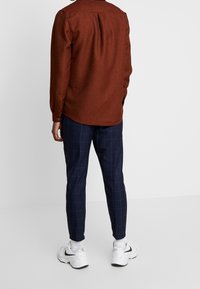 Man som står med ryggen mot kameran, iförd en rostfärgad långärmad skjorta, marinblå rutiga byxor, vita strumpor och vita sneakers med svart logotyp.