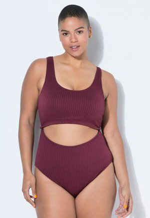 Femme aux cheveux courts portant un maillot de bain une pièce bordeaux côtelé avec découpes, debout devant un fond blanc.
